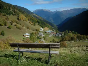 Alpenlandschaften der Savoie - Holzbank mit Blick auf die Alm, die Häuser eines Dorfes, die Bäume im Herbst, die Wälder und die Berge