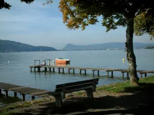 Annecy lake - Shore with a bench and a tree, lake, wooden pontoons, boat and hills in background