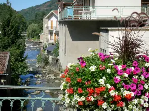 Ax-les-Thermes - Kurort: mit Blumen geschmückte Brücke überspannend den Fluss, Häuser und Bäume am Wasserrand