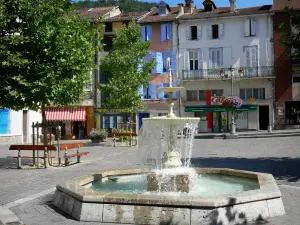 Ax-les-Thermes - Kurort: Brunnen des Platzes Roussel, Bäume, Sitzbank und Häuserfassaden