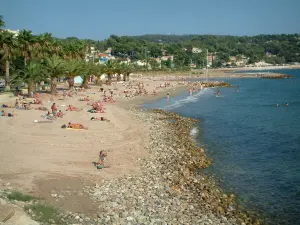 Bandol - Le palme, spiaggia sabbiosa del resort con i turisti, ciottoli, Mar Mediterraneo, case e foreste in background