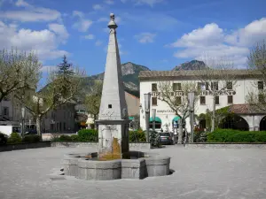 Castellane - Fountain, trees and houses of the Marcel Sauvaire square; clouds in the blue sky