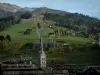 La Clusaz - Church bell tower and chalets of the winter and summer sports resort, alpine pastures (high meadows), ski area with ski lifts and trees in autumn