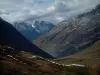 Les cols de la route des Grandes Alpes - Col de la Croix-de-Fer: De la route de la Croix-de-Fer, vue sur des alpages (hauts pâturages), un lac et des montagnes aux cimes recouvertes de neige avec un ciel nuageux (route des Grandes Alpes)