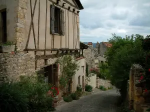 Cordes-sur-Ciel - Calle en cuesta empedrada, con flores, plantas, casas de estructura de madera y cielo nublado
