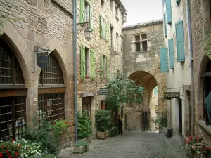 Cordes-sur-Ciel - Portal Peint (befestigtes Tor) das das Geschichts- und Kunstmuseum Charles-Portal birgt,  gepflasterte Strasse geschmückt mit Blumen und Pflanzen, Häuser aus Stein mit Fassaden dekoriert  mit Schildern aus Schmiedeeisen und Kunstgalerien