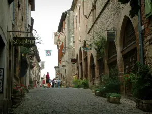 Cordes-sur-Ciel - Rue pavée en pente bordée de fleurs et de plantes, maisons en pierre aux façades ornées d'enseignes en fer forgé et de drapeaux, échoppes et galeries d'art