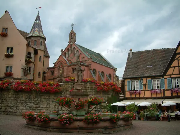 Eguisheim - Flower-bedecked square (geraniums) with its fountain, the Eguisheim castle, the Saint-Léon-IX chapel, a house and a café terrace