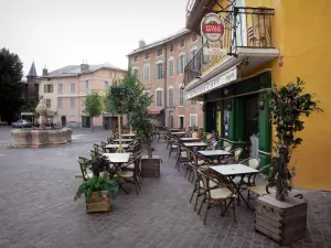 Embrun - Place Barthelon : terrasse de café, fontaine et maisons aux façades colorées de la vieille ville