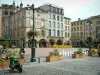 Épinal - Place des Vosges avec plantes, fleurs, jets d'eau, terrasses de cafés et maisons à arcades