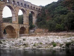 Gard Brücke - Dreistöckige römische Aquäduktbrücke mit Bogen (Bogenbrücke, antikes Bauwerk), Ufer bepflanzt mit Bäumen und Fluss Gardon; auf der Gemeinde Vers-Pont-du-Gard