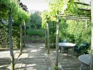 Gärten des Priorats Notre-Dame d'Orsan - Tische und Stühle unter der Pergola, Pflanzen und Bäume im Hintergrund