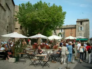 Gordes - Square in the village with café terraces, parasols, trees and houses