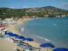 Les Issambres - Parasols and sunbeds with tourists, sandy beach of the seaside resort, the Mediterranean Sea with yellow buoys, villas (Provencal houses) on the hill