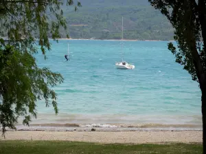 Lac de Sainte-Croix - Plage de Sainte-Croix-du-Verdon, retenue d'eau couleur émeraude, bateau, planche à voile et colline couverte d'arbres ; dans le Parc Naturel Régional du Verdon