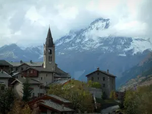 Lanslevillard - Kerktoren en huizen in het dorp, besneeuwde bergen op de achtergrond en bewolkte hemel, in de Haute-Maurienne (perifere zone van de Vanoise National Park)
