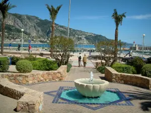 Menton - Small fountain decorated with blue mosaics, palm trees, port and mountain in background