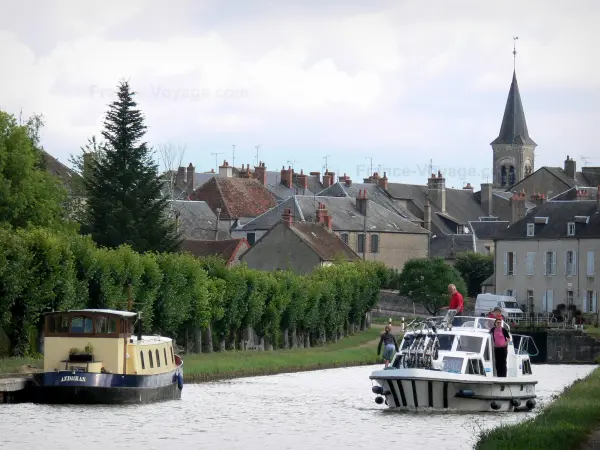 Nivernais-Kanal - Motorjacht fahrend auf dem Nivernais-Kanal, in einer Reihe stehende Bäume, Kirchturm der Kirche Saint-Jean-Baptiste, und Häuser des Dorfes Châtillon-en-Bazois