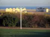 Ouistreham-Riva-Bella - Acera con una luz de la calle, césped, arbustos, casetas de playa y el mar (Canal Inglés) en el fondo