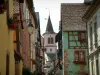 Riquewihr - Maisons colorées aux façades ornées d'enseignes en fer forgé et aux fenêtres fleuries de géraniums, clocher de l'église en arrière-plan