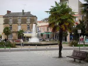 Les Sables-d'Olonne - Bench, palm tree in foreground, square with a fountain and rosebushes (roses), and houses of the town centre