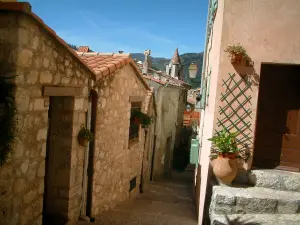Sainte-Agnès - Calle estrecha asfaltada, escalera de piedra, maceteros y torre de la iglesia