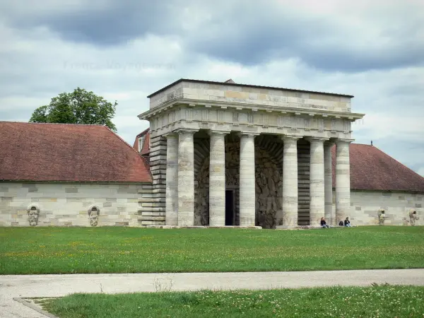 Saline royale d'Arc-et-Senans - Bâtiment des Gardes (entrée de l'ancienne saline) avec ses colonnes ; pelouses parsemées de fleurs, allée et ciel orageux