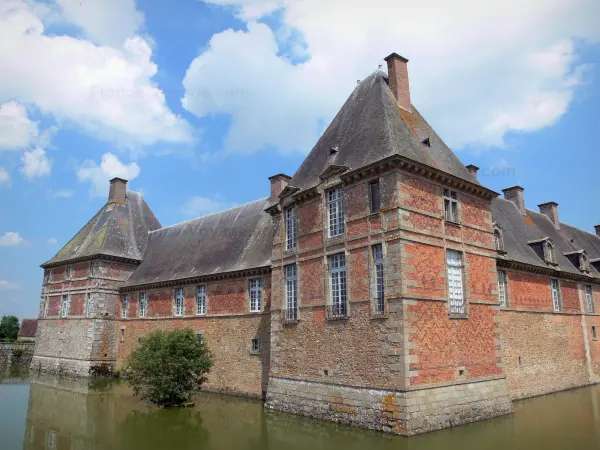 Schloss von Carrouges - Schloss aus Backstein und Stein umgeben von Wassergräben, und Wolken im blauen Himmel; im Regionalen Naturpark Normandie-Maine