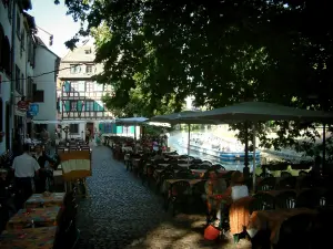 Strasbourg - La Petite France (ancien quartier des tanneurs, meuniers et pêcheurs) : terrasses de restaurants ombragées par un arbre et rivière (l'Ill) avec un bateau