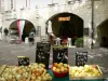 Uzès - Place aux Herbes: stand di frutta e verdura del mercato in primo piano, terrazza del ristorante, negozio e casa a portici (archi)