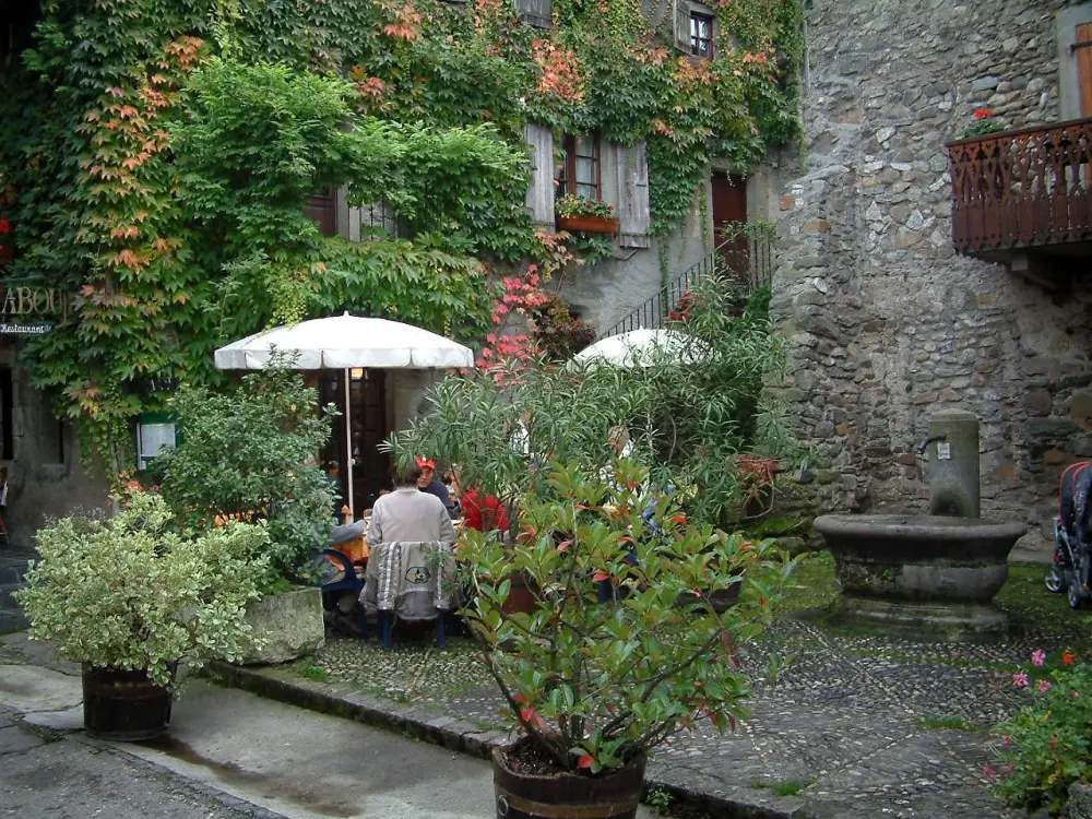 Yvoire - Petite place pavée avec fontaine, terrasse de restaurant, arbustes en pots et façade couverte de lierre