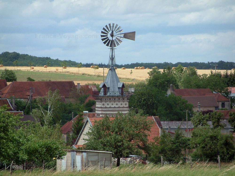 Guide de l'Aube - Paysages de l'Aube - Éolienne, arbres et maisons d'un village, champ avec des bottes de paille en arrière-plan