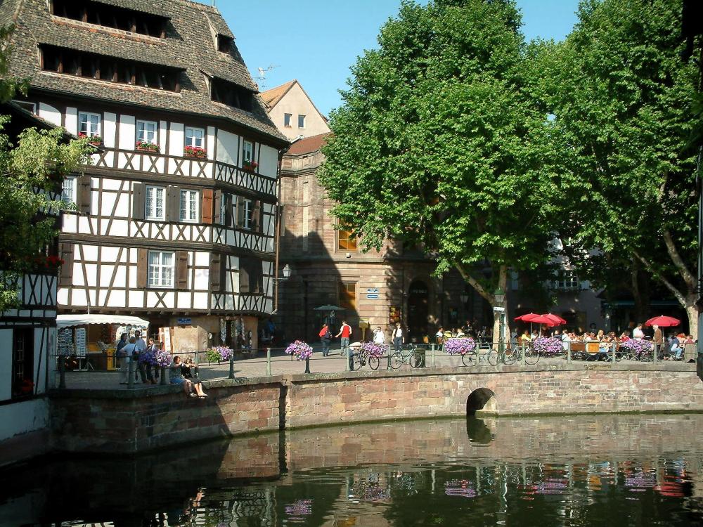 Guide du Bas-Rhin - Strasbourg - La Petite France (ancien quartier des tanneurs, meuniers et pêcheurs) : rivière (l'Ill), berge fleurie avec boutique, terrasse de café, arbres et maisons blanche à colombages au toit pentu