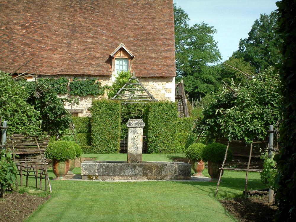 Guide du Centre-Val de Loire - Jardins du prieuré Notre-Dame d'Orsan - Jardin du cloître d'inspiration médiévale avec fontaine aux quatre jets, plantes, arbres et ancien monastère en arrière-plan