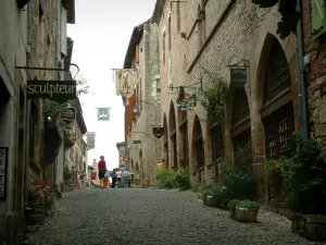 Cordes-sur-Ciel - Rua inclinada pavimentada forrada com flores e plantas, casas de pedra com fachadas adornadas com placas e bandeiras de ferro forjado, barracas e galerias de arte