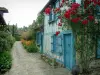 Gerberoy - Blue house and its climbing rosebush (red), narrow paved street lined with flowers and plants