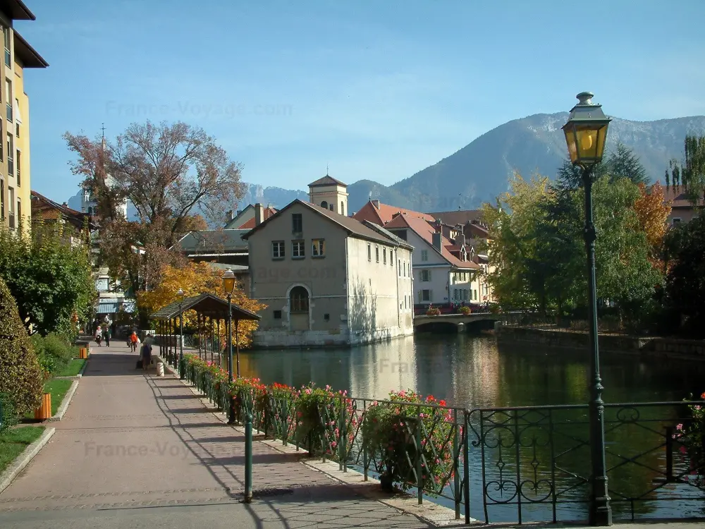 Guide of the Haute-Savoie - Annecy - Flower-bedecked bank, lampposts, trees, Thiou canal, houses of the old town along the water and mountains in background