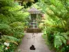 Hell-Bourg - Jardin créole de la maison Folio : chat dans l'allée d'entrée bordée de végétation tropicale, avec vue sur la fontaine des Trois Grâces et le guétali (kiosque)