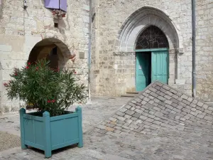 Lauzerte - Corner of the Place des Cornières square, portal of the Saint-Barthélemy church, arcaded house and potted blooming shrub