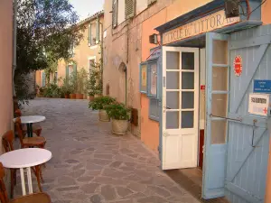 Ramatuelle - Ruelle du village avec tables et chaises d'un bar, plantes et fleurs en pots et maisons aux façades colorées