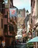 Ribeauvillé - Rue avec drapeau suspendu, maisons colorées aux fenêtres ornées de fleurs et tour des Bouchers (ancien beffroi) en arrière-plan