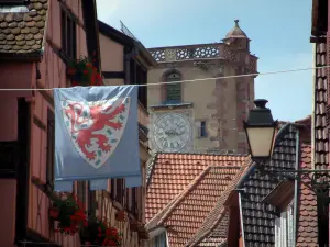 Ribeauvillé - Aufgehängte Fahnen, farbige Fachwerkhäuser, Fenster geschmückt mit Geranien, Hausdächer und Turm Boucher (ehemaliger Wachturm) im Hintergrund