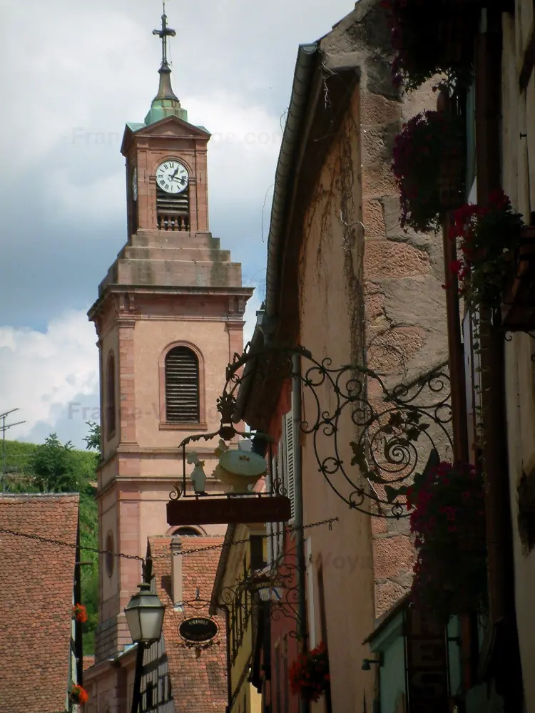 Riquewihr - Gekleurde huizen met gevels versierd met smeedijzeren tekenen en kerktoren op de achtergrond