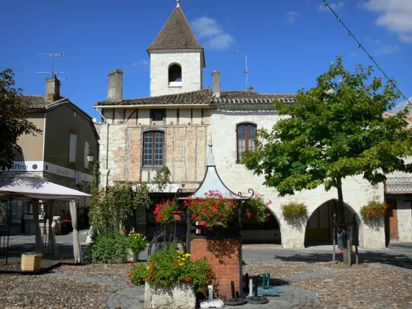 Tournon-d'Agenais - Bastide: blumengeschmückter Brunnen (Blumen) und Häuser der Platzes, Glockenturm überragend die Gesamtheit