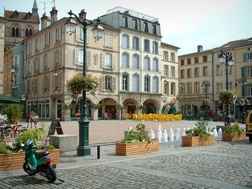 Guide des Vosges - Épinal - Place des Vosges avec plantes, fleurs, jets d'eau, terrasses de cafés et maisons à arcades